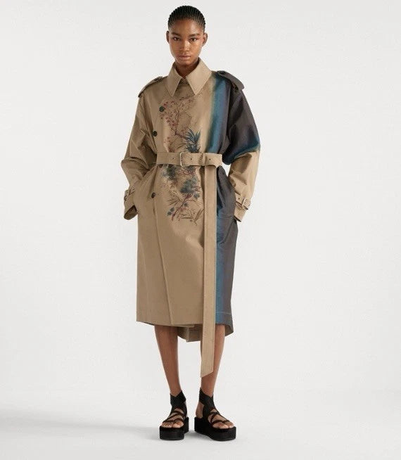 A model wearing a beige trench coat with blue and black color-blocked panels and floral embroidery, paired with black platform sandals, against a plain white studio background.