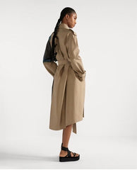 A model shown from the back wearing a mid-length beige trench coat with a belted waist, paired with black platform sandals and a dark backpack, against a plain light gray studio background.