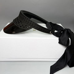 A black visor hat with a textured woven pattern band, a glossy black brim, and a long black ribbon tie, displayed on top of a white box.