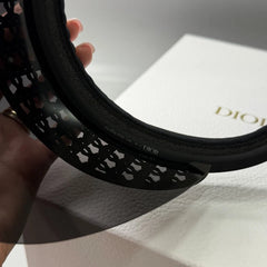 A close-up of a black Dior hat featuring an intricate geometric lattice cutout pattern on the brim, held by a hand over a white Dior-branded box.