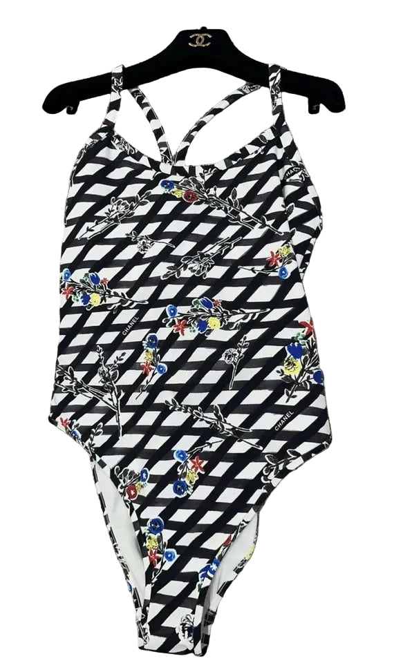 A one-piece swimsuit with a black and white geometric pattern and colorful floral accents, hanging on a black hanger with a Chanel logo, placed on a textured gray fabric surface.