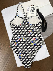 A one-piece swimsuit with a black and white geometric diamond pattern and colorful floral print, laid flat on white Chanel packaging on a wooden floor.