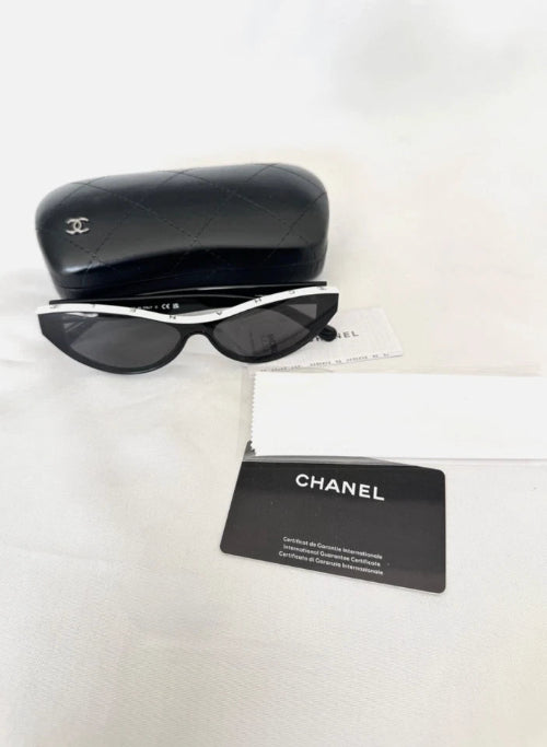 Chanel sunglasses with a black and white frame, shown with a black quilted case, a white box, and a black Chanel card on a white fabric background.