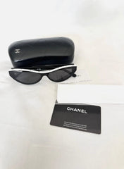 Chanel sunglasses with a black and white frame, shown with a black quilted case, a white box, and a black Chanel card on a white fabric background.