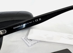 Close-up of a black Chanel sunglasses arm with white printed branding and text, resting on a white textured surface with a white dust bag and part of a black quilted case visible.