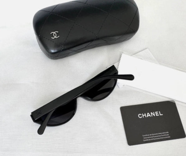 Black Chanel sunglasses with a matching black quilted leather case, shown on a white fabric background with a white dust bag and a Chanel card.