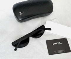 Black Chanel sunglasses with a matching black quilted leather case, shown on a white fabric background with a white dust bag and a Chanel card.