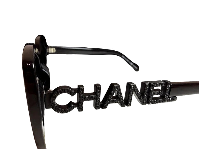 A pair of black Chanel sunglasses with a prominent silver-tone CHANEL logo on the frame, displayed next to its black branded box on a light marble surface.