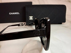 A pair of black Chanel sunglasses with the iconic interlocking CC logo on the temple, displayed next to its black presentation box on a light-colored surface.