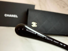 Close-up of a black Chanel eyeglass arm with white printed model text, shown in front of a black Chanel box and matching black quilted pouch on a light marble surface.