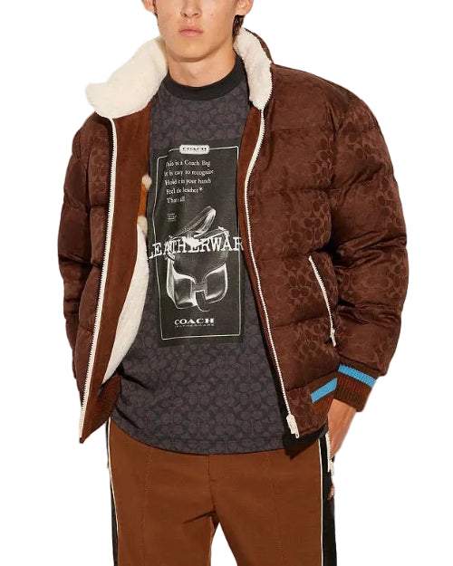 Person wearing a brown patterned jacket with white lining, a dark t-shirt with graphic design, and brown pants on a light gray background.