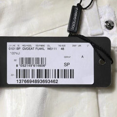 A close-up of a white garment tag attached with a black string to a black Dolce & Gabbana brand tag, showing product details and barcodes on a white fabric background.