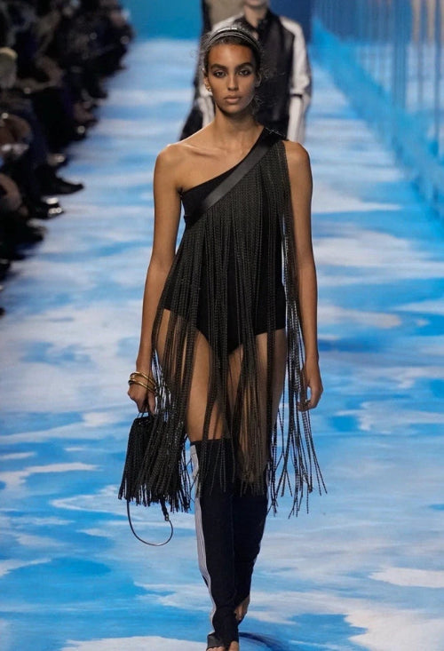 A model walks a fashion runway wearing a black fringe dress with a sheer mesh overlay and a black handbag.