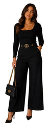 A woman modeling a black long-sleeve jumpsuit with a square neckline, wide legs, and a gold double-G buckle belt. She is holding a black quilted Gucci handbag with a gold chain strap in an indoor setting.