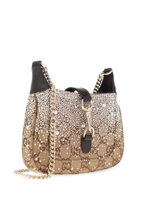 A Gucci shoulder bag covered in sparkling rhinestones and monogrammed with gold-tone hardware, featuring a gold chain strap and black leather trim, set against a plain white background.