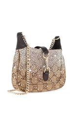 A Gucci shoulder bag covered in sparkling rhinestones and monogrammed with gold-tone hardware, featuring a gold chain strap and black leather trim, set against a plain white background.