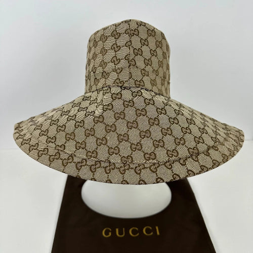 A Gucci wide-brim sun hat in beige and brown GG Supreme canvas, displayed on a white mannequin head with a dark brown Gucci dust bag underneath.
