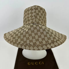 A Gucci wide-brim sun hat in beige and brown GG Supreme canvas, displayed on a white mannequin head with a dark brown Gucci dust bag underneath.