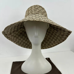 A wide-brimmed sun hat with a beige and brown repeating monogram pattern, displayed on a white mannequin head. The hat features a Gucci-style GG monogram pattern, and a dark brown fabric pouch is visible at the bottom.