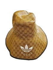 A Gucci x Adidas bucket hat in the signature brown GG monogram canvas, featuring a white Adidas trefoil logo and the word 'GUCCI' on the front, resting on a beige carpet.