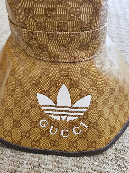 A close-up of a Gucci-branded sun visor featuring the classic brown GG Supreme monogram pattern on a tan background, with a white Adidas trefoil logo and 'GUCCI' text on the front.