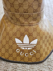 A close-up of a Gucci-branded sun visor featuring the classic brown GG Supreme monogram pattern on a tan background, with a white Adidas trefoil logo and 'GUCCI' text on the front.