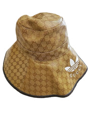 A bucket hat featuring the brown GG monogram pattern on a tan background, with a white Adidas trefoil logo and 'GUCCI' text on the brim, resting on a beige carpet.