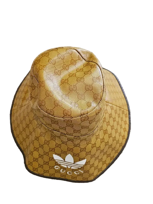 A Gucci x Adidas bucket hat in the signature GG Supreme canvas, featuring a mustard yellow and brown pattern with a white Adidas trefoil logo and Gucci branding on the brim, displayed on a beige carpet.