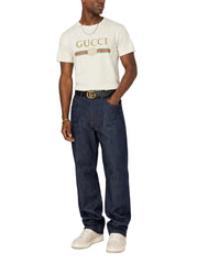 A man modeling a cream-colored Gucci t-shirt with the brand's signature green and red stripe logo, paired with dark blue jeans, a black belt with a gold buckle, and white sneakers, against a plain light gray background.