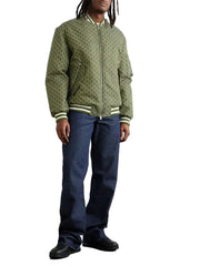 A man wearing an olive green zip-up jacket, dark blue jeans, and black shoes, standing against a plain off-white wall.