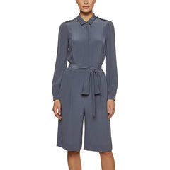A woman modeling a slate blue long-sleeve collared shirt-jumpsuit with a matching tie belt at the waist, shown against a plain light beige studio background.