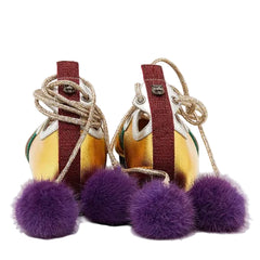 A pair of gold sneakers with burgundy straps, chunky metallic beige laces, and large purple faux-fur pom-poms hanging from the sides, photographed on a light gray studio background.