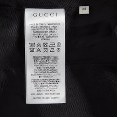 A close-up of a white Gucci garment care label sewn onto black textured fabric, with the Gucci brand name clearly printed at the top and a small size marker reading 38 nearby.