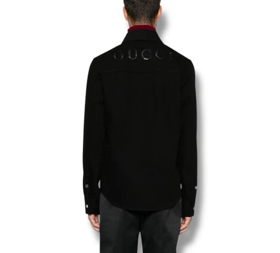 A man viewed from the back wearing a black Gucci shirt jacket with silver rhinestone lettering across the upper back, paired with dark trousers.