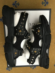 Black studded sandals on a white box with 'gucci' branding against a patterned brown background.