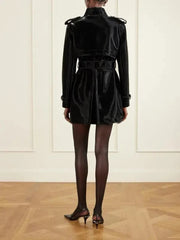 A model wearing a black leather trench coat, black tights, and black high heels, standing in a minimalist room with a light-colored wall and wooden floor.