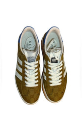 A pair of Gucci GG Marmont low-top sneakers in olive brown suede with white laces, white side stripes, and blue accents on the collar and heel, shown from a top-down view on a white background.