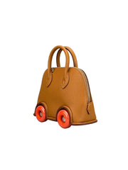 A small structured handbag designed like a toy car, made of tan textured leather with bright orange wheels and gold-tone hardware, photographed on a neutral gray studio background.