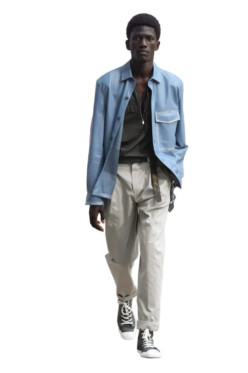 Man wearing a light blue shirt over a dark sweater with beige pants and black shoes on a white background