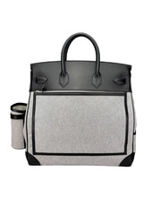 A structured tote bag with a light gray heathered front panel, black leather top and handles, and black trim, shown on a neutral gray background. A matching insulated cup is partially visible to the left.