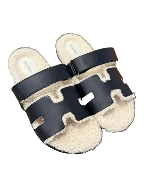 Pair of slippers with black straps and white fur lining on a wooden floor.