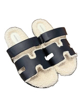 Pair of slippers with black straps and white fur lining on a wooden floor.