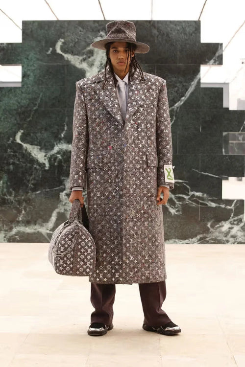 Man in a patterned coat and hat holding a suitcase against a marble wall.