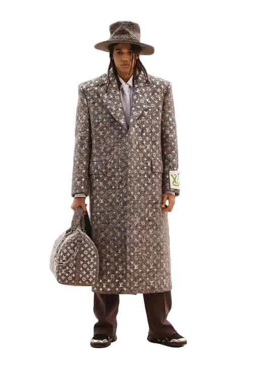 Man in a patterned coat and hat holding a suitcase against a marble wall.