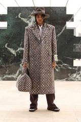 Man in a patterned coat and hat holding a suitcase against a marble wall.