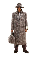 Man in a patterned coat and hat holding a suitcase against a marble wall.