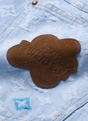 Close-up of light blue denim jeans featuring a brown suede cloud patch with embossed Louis Vuitton branding, a blue embroidered monogram motif, and a metal button at the top.