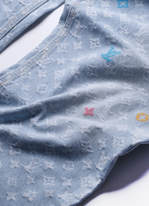 A close-up of a light blue denim garment featuring an all-over embossed Louis Vuitton monogram pattern and small embroidered LV logos in blue, pink, and yellow.