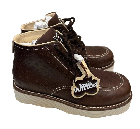A pair of Louis Vuitton dark brown embossed leather ankle boots with white contrast stitching and a cream-colored sole, displayed on a white textured surface against a gray wall.