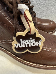 A close-up of a brown leather Louis Vuitton shoe with a white and gold paper tag attached to the side, showing the brand name 'Louis Vuitton' in gold lettering.